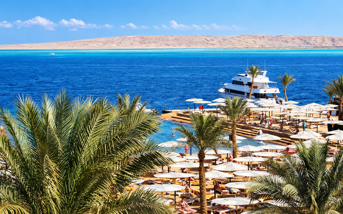 A resort beach with palm trees, umbrellas and a yacht offshore