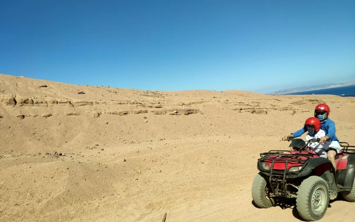 Two helmeted riders on a quad beside a desert coast
